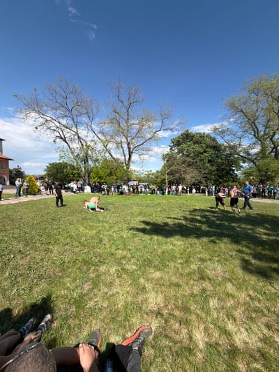 The image depicts an outdoor scene, possibly in a park or grassy area in the city of Dragomir, Bulgaria. People are gathered, appearing to observe something taking place on the grass, where at least one individual is lying down, wearing green clothing. A line of spectators can be seen in the background, along the edge of the grass. Trees provide shade and frame the scene, with a clear blue sky visible above. The buildings appear at the very edge of the image, suggesting the edge of the populated area or town.