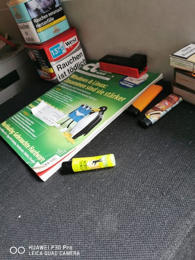 The image displays a collection of items on a dark, textured surface. In the background, partially visible, are two cigarette packs, one blue and white with "Pueblo" branding, and another red and white with "West" branding, both bearing warning labels in German. A black and red cigarette rolling machine rests on top of the "West" pack.

Dominating the foreground is a stack of magazines, predominantly green with headlines in German. One magazine's cover features cartoon penguins and text that includes "Windows & Linux" and "Zusammen sind sie stärker". A folded white slip of paper is attached to the magazine.

In front of the magazines, two lighters are placed. The first is orange, and the second is decorated with various images. A yellow object, possibly a highlighter or marker, with black text is positioned near the bottom of the frame.

The bottom of the image contains a watermark that reads "HUAWEI P30 Pro LEICA QUAD CAMERA".

No people are present in the image. The scene appears to be an indoor still life arrangement. The specific location in Bad Oeynhausen, Germany, cannot be determined from the visual information provided.