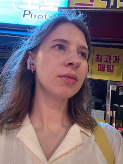 A light-skinned woman with light brown hair is pictured from the chest up, facing to the right. She is wearing a light-colored collared shirt and a yellow strap across her shoulder. She has on purple earbuds and a small hoop earring in her visible ear. The background appears to be an outdoor urban setting at night, with illuminated signs. One sign in the upper left corner displays the word "Photo" in large, bright blue letters against a blue background. Another sign in the upper right features Korean text in red lettering on a yellow background, including "최고가" and "매입". The overall impression is a selfie taken in a city.