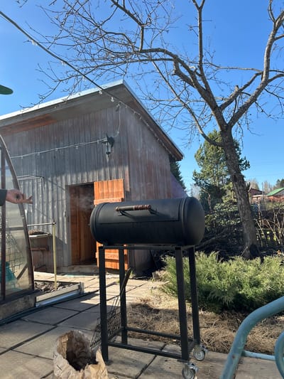The image depicts an outdoor scene with a black cylindrical smoker situated on a black metal frame with wheels, resting on a paved surface next to a wooden building. The building has a window or opening through which smoke is visible. A bare tree branch is visible in the upper left corner against a clear blue sky. Part of a greenhouse is seen in the foreground. A bag is positioned on the paved surface to the right of the smoker. A partially visible human hand is in the foreground, giving the peace sign. The location is specified as Bashur, Russia.