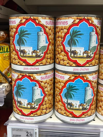 The image presents a close-up view of four cylindrical metal cans of chickpeas, specifically "pois chiches," displayed on a white metal supermarket shelf. Two cans are stacked on top of two others. Each can bears an identical label design: a light brown background patterned with chickpeas, framing a central red-bordered quatrefoil-shaped vignette. This vignette illustrates an arid landscape featuring a green palm tree on the left, two to three brown camels (one standing, one resting), and a white, conical-roofed tower with adjacent white buildings under a clear blue sky. Red text reading "sultanines" is positioned above the vignette, and "pois chiches" below it. A partial view of a fifth can is visible to the right. To the left, a portion of a yellow and green packaged food item is partially obscured by a white plastic shelf divider. A white digital price tag affixed to the edge of the shelf below the cans displays the text "POIS CHICHES SULTAM DAUCY 530G," a price of "2.10 €," and a barcode. The scene is an indoor retail environment consistent with a supermarket in France, based on the French labeling and Euro currency, but the specific city of La Valette-du-Var cannot be determined from the visual information provided. No individuals are present in the image.