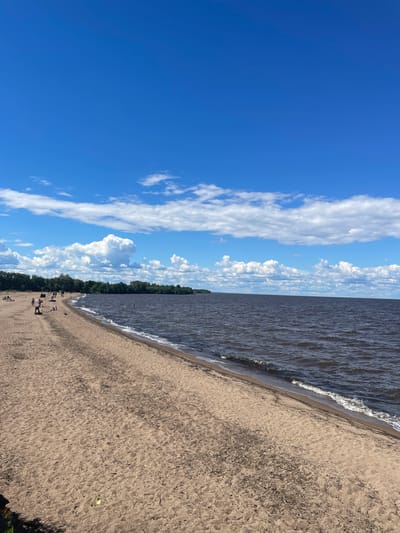 The image shows a sandy beach on the shore of a large body of water, likely Lake Beloye, in Belozersk, Russia. The sky is bright blue with scattered white cumulus clouds. The water is dark and choppy with small waves breaking on the shore. A line of green trees borders the beach in the distance. There are several people on the beach, some sunbathing, others walking or sitting. A few people are in the water. The sand is light brown and appears to be a mixture of sand and small debris. The overall scene depicts a warm, sunny day at the beach.