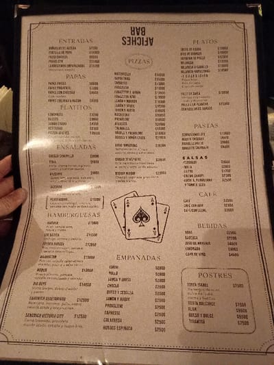 The image shows a menu from a restaurant called "Afliches Bar" in Victoria, Argentina. The menu is written in Spanish and lists a variety of food and drink options, including pizzas, pastas, salads, empanadas, sandwiches, desserts, and drinks. The restaurant is located at coordinates -34.452 latitude and -58.54 longitude. There is a pair of playing cards, the Ace of Spades, displayed at the top of the menu.  The image is taken from an angle looking directly at the menu. The image is a close-up of the menu and shows no people.