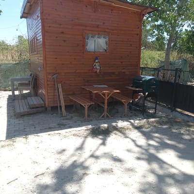 A wooden cabin stands to the left of the frame, its walls constructed from vertical planks of reddish-brown wood. A small window with a floral patterned curtain is visible on the cabin's side. To the right of the window, a decorative doll with a blue hat and colorful clothing is attached to the wall. In front of the cabin, a wooden picnic table with benches is set up. Behind the table, a green barbecue grill is positioned under a dark metal structure. The ground around the cabin is a mixture of light-colored sand and some patches of grass, with a cobblestone path to the right. The scene is shaded by dappled sunlight filtering through unseen trees. In the background, the foliage of trees and what appears to be a green tarp are visible. The overall setting suggests an outdoor recreational area or a backyard. The lighting indicates it is daytime, likely a sunny day. No visible text is present.