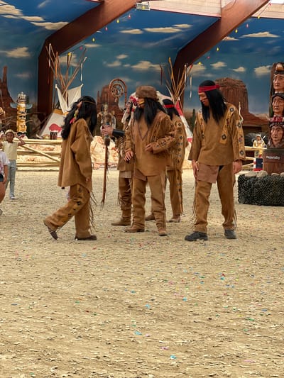 This image depicts an outdoor performance with individuals dressed as Native Americans. The scene appears to be set in a themed park or attraction, possibly related to a historical or cultural reenactment. In the foreground, three people in traditional Native American attire are the primary subjects. They are wearing brown leather outfits with fringes and decorative patterns. The person on the left is a child, facing left and walking away from the camera. The two individuals to their right are adults. One adult is facing forward and slightly to the left, while the other is facing to the right and looking down. A fourth person, also in Native American attire, is partially visible behind the first adult, facing left.

The background features a painted mural of a desert landscape with mesas and a cloudy sky. There are also several props, including teepees, totem poles, and a dreamcatcher. A wooden fence runs across the middle ground. In the far background, a structure with a wooden roof and hanging lights is visible. The ground is covered in sand or dirt, with scattered confetti. On the right side of the image, a wooden barrel with the text "PonyParkCity" is visible, suggesting the location is PonyParkCity in Collendoorn, Netherlands. Other figures, including what appear to be mannequins or statues of Native Americans and a small statue of a horse with a rider, are also present in the background. The overall atmosphere suggests a lively event or attraction.
