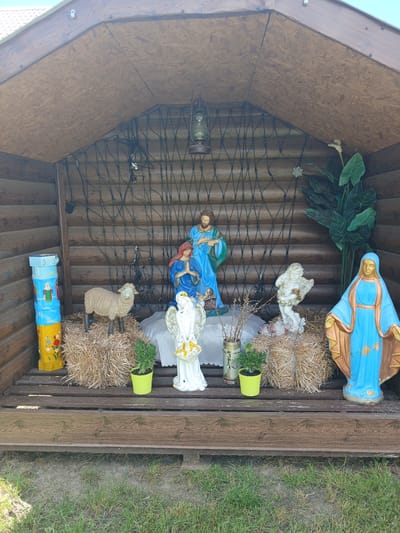 The image shows an outdoor nativity scene set within a wooden structure. The scene includes statues of Mary, Joseph, and an infant Jesus. There is also a statue of an angel, a lamb, and a large statue of Mary to the right of the scene. The figures are positioned on hay bales and a wooden platform. In the background, there is a wire mesh with lights draped across it, and a hanging lantern. To the left of the scene, there is a tall, colorful cylindrical object with painted religious imagery. Green potted plants are placed around the nativity figures. The structure itself is made of logs, and the ground in front is covered with grass. The image was taken in Doslidnytske, Ukraine.