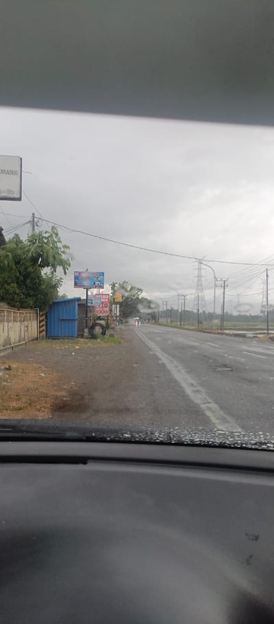 The image captures a view from inside a vehicle, looking out onto a wet asphalt road under an overcast sky. Water droplets cling to the windshield in the foreground, indicating recent or ongoing rain. The time of day appears to be daytime, but the light is dim and grey.
On the left side of the road, there's a dirt and gravel shoulder beside a concrete fence. Behind the fence, green trees are visible. A blue corrugated metal shack stands next to the road, near several billboards. Further down the road, power lines and utility poles line the left side.
On the right side, the road stretches into the distance, appearing slick with moisture. A broken white line marks its center. In the mid-distance, a few vehicles are discernible with their headlights on. Large electricity transmission towers rise into the cloudy sky in the background, contrasting with what appear to be flat fields or rice paddies extending to the horizon on the right.
Visible text on signs includes "MANE" and "G.Go" (likely referring to Glee Gogo, Indonesia) on a white billboard in the upper left. A prominent blue billboard advertises "K*** MANDIRI" and "ALUMINIUM," with another sign beneath it displaying "RAJA ALUMINIUM." The scene portrays a semi-rural road environment in Glee Gogo, Indonesia, during gloomy, wet weather.