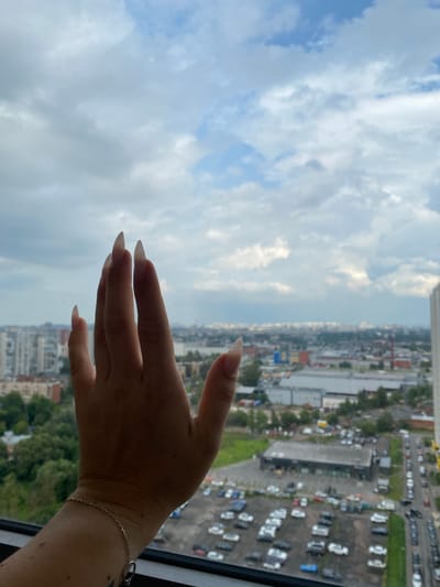 The image shows a close-up of a person's hand, with long, pointed fingernails, held up against a window. The hand is casting a shadow on the glass. Through the window, a cityscape is visible, including buildings, trees, and a parking lot filled with cars. The sky is cloudy and overcast. The location is identified as Ostrovki, Russia. The image appears to be taken from a high vantage point, possibly a high-rise building, looking out over the urban landscape. The overall tone is neutral, with no specific emotional cues.