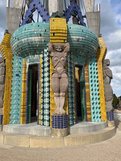 The image shows a detailed close-up of a colorful, art deco-inspired sculpture. The sculpture features a recumbent nude figure carved from stone, with stylized musculature and a serene expression. This figure is positioned atop a layered structure adorned with vibrant ceramic tiles in shades of turquoise, yellow, and blue, interspersed with some black and white tiles. The tiled sections create geometric patterns, including wave-like motifs and rectangular arrangements.
Beneath the recumbent figure, another stone carving of a reclining person is partially visible, suggesting a continuation of the theme. The sculpture is integrated with concrete architectural elements, forming tiered sections and arches. In the background, glimpses of a cloudy sky and some greenery can be seen, indicating an outdoor setting. The lighting suggests it is daytime, possibly late afternoon or early morning given the cloud cover. The overall impression is one of a significant public art installation, likely a fountain or monument, with intricate craftsmanship and a bold aesthetic. The image is oriented vertically, with the sculpture appearing to rise upwards.