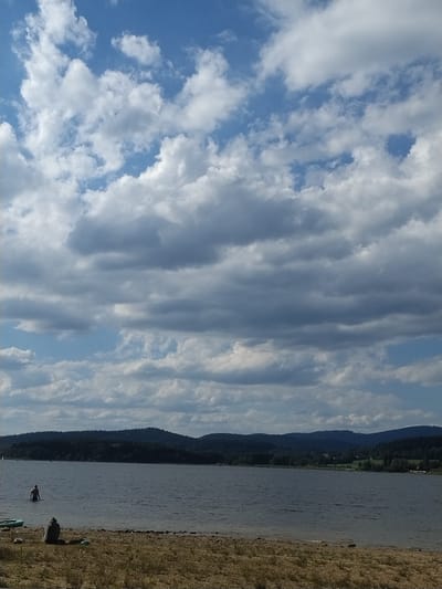 The image captures a serene lake scene with a backdrop of rolling, forested hills under a partly cloudy sky. The foreground features a sandy shoreline with dry grass and sparse vegetation. A dark, irregularly shaped rock sits on the shore, accompanied by a blue inflatable object and what appears to be a green bag or tarp. In the water, a solitary person stands knee-deep, facing away from the camera. Further out on the lake, distant sailboats can be seen, and the water is calm with gentle ripples. The overall impression is one of a quiet, natural setting, possibly in Radslav, Czechia, on a pleasant day.