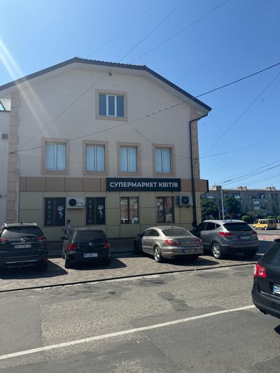 The image shows a storefront in Sarny, Ukraine, labeled "СУПЕРМАРКЕТ КВІТІВ" (Flower Supermarket). The building is light beige with a dark grey roofline and tan-colored trim around the windows and lower facade. There are multiple windows on the upper and lower floors. The lower facade features two large windows, each with vertical text that reads "КВІТИ" (Flowers) and a stylized rose graphic. An outdoor air conditioning unit is mounted between these windows.
Several cars are parked in front of the building. On the left, a black Nissan Rogue with license plate BK 6106 CE is visible. Next to it is a black Volkswagen Golf with license plate BK 6982 IT. Further to the right, a silver Volkswagen CC with license plate BK 0389 IX is parked, followed by a grey Hyundai Tucson with license plate BK 3077 CO. A person in a light-colored shirt is standing near the air conditioning unit, holding a smartphone. In the background, apartment buildings and a yellow taxi are visible, suggesting an urban or suburban setting. The sky is clear and blue.