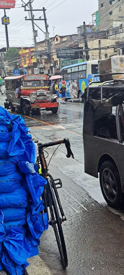 The image depicts a street scene in Mandaluyong, Philippines, during a rain shower. A red and silver jeepney, a common mode of public transportation in the Philippines, is parked on the wet asphalt road. Beside it, a stack of large blue tarpaulin-covered bundles is leaning against a wall. A bicycle with a brown handlebar is positioned in front of the bundles, its front wheel partially visible.

In the background, several buildings line the street, some with colorful facades and numerous wires strung overhead, indicative of a bustling urban environment. A blue bus with graphics on its side is also visible, suggesting ongoing traffic. Two young individuals, possibly students in uniforms with yellow shirts and blue skirts, are walking on the wet sidewalk, one holding a pink umbrella. The overall atmosphere is that of a typical day in a Philippine city, albeit with the addition of rain. The sky appears overcast, and water is visible on the road surface, reflecting the surrounding objects.