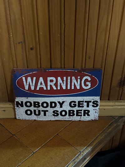 The image shows a weathered, rectangular metal sign mounted on a wood-paneled wall. The sign features the word "WARNING" vertically aligned in a red oval against a blue background on the left side. To the right, in black block letters against a white background, is the phrase "NOBODY GETS OUT SOBER." The sign exhibits signs of rust and wear. The surrounding environment consists of wooden paneling to the left and a vertical light wood trim piece to the right. The presence of tiled surfaces further to the right suggests the sign is indoors. This image doesn't indicate the location is in Lilkovo, Bulgaria.