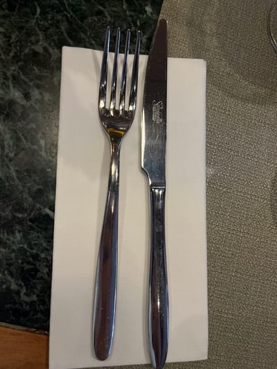 A polished stainless steel four-tine dinner fork is positioned to the left of a polished stainless steel dinner knife. Both utensils rest horizontally aligned on a folded white fabric napkin. The knife blade is etched with "Salvinelli" above "INOX FORGE". The napkin covers part of two distinct table surfaces: a dark green marble-patterned surface on the left and a light beige, textured surface (possibly a placemat or tablecloth) on the right. A small segment of a brown wooden surface is visible in the bottom-left corner of the frame. The scene is illuminated by an overhead light source, causing reflections on the metallic cutlery. No people or actions are depicted. The image contains no discernible features, landmarks, or contextual elements that would allow for the identification of a specific location within the city of Baarle-Nassau, Netherlands.
