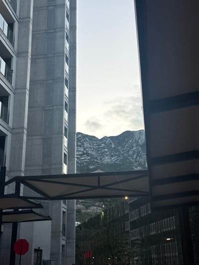 This image depicts a scene in Santa Catarina, Mexico, looking through an urban architectural corridor towards a mountainous background. On the left, a multi-story building with a light gray facade features numerous balconies with glass railings and vertical louvered sections. A dark, vertical structural element partially frames the right side of the image. A modern pedestrian walkway, composed of a dark metal frame and a light-colored fabric canopy, spans horizontally across the lower mid-ground. Below this walkway, ground-level structures, dark foliage, and faint red cylindrical objects are discernible. The background is dominated by a prominent mountain range, characterized by rugged, rocky slopes and visible vegetation, situated under a light, partly cloudy sky. No individuals are present in the scene.