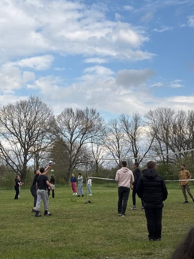 The image shows a group of people playing volleyball in a grassy field. A volleyball net is visible in the center of the frame, with bare trees in the background. The sky is visible above the trees and is blue with white clouds. There are approximately 12 people in the scene, wearing various casual clothes. Some people are actively engaged in playing volleyball, with at least one person hitting the ball. Others are observing the game. The specific location in Gromshin, Bulgaria, is not identifiable from the image. The environment appears to be a park or open recreational space.