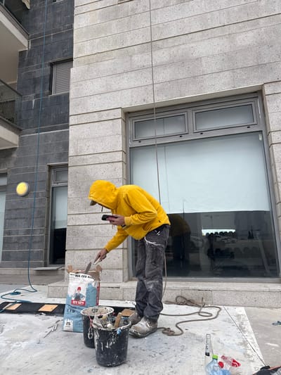 The scene depicts an outdoor ground-level area adjacent to a modern multi-story building in Tirat HaCarmel, Israel. The building features two distinct facades: the left side comprises dark grey horizontally-laid rectangular tiles with a balcony and a circular yellow wall-mounted light fixture, while the right side is constructed of lighter grey horizontally-laid rectangular tiles with large, dark-framed windows.
A single individual, a male, is present, wearing a bright yellow hooded sweatshirt, dark, paint-splattered work pants, and light-colored work boots. His hood is up, partially obscuring his face, though a dark cap is visible underneath. He is bent forward, actively scooping material from an open white and blue bag, labeled in Hebrew with "Nirlat Acrylic Plaster" and "25 kg", using a flat trowel held in his right hand. In his left hand, he holds a dark electronic device, possibly a mobile phone.
On the concrete ground around the individual are two black plastic buckets containing various tools and residual materials, a coiled rope, and scattered fragments of dried plaster or construction debris. In the lower right foreground, a plastic water bottle, a smaller white plastic bottle, and a red plastic bag are discarded. Two ropes descend from the upper sections of the building: a blue rope along the darker facade on the left, and a thinner, dark rope along the lighter facade, passing behind the individual. The large window on the right facade reflects a partial interior view, including a light-colored roller blind, and a blurred exterior reflection of what appears to be a street or parking area with vehicles.