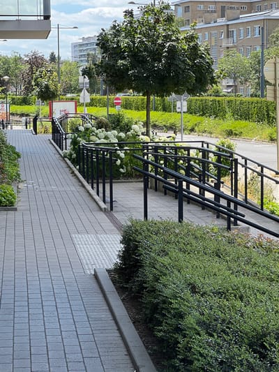 The image shows a paved walkway with a ramp and handrails in Evere, Belgium. The walkway is constructed of grey rectangular pavers. A tactile paving surface is visible on the walkway. Green hedges and flowering plants, including white hydrangeas, line the walkway and ramp. In the background, there are buildings, trees, and streetlights. A traffic sign with a red circle and white horizontal bar, indicating "no entry," is visible. A modern building with multiple floors and balconies is prominent in the background. The sky is partly cloudy. No people are visible in the image.