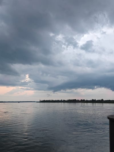 A wide shot captures a body of water under a heavily clouded sky. The sky is filled with dark, imposing storm clouds, with a few lighter patches hinting at the sky beyond. The water's surface is rippled, reflecting the muted tones of the sky. In the distance, a thin strip of land is visible, densely forested with trees. Two small boats are present on the water: one closer to the left bank, and another further out, near the right bank. A dark railing or edge of a structure is visible in the lower right corner of the image. The location is identified as Kazachinskoye, Russia.