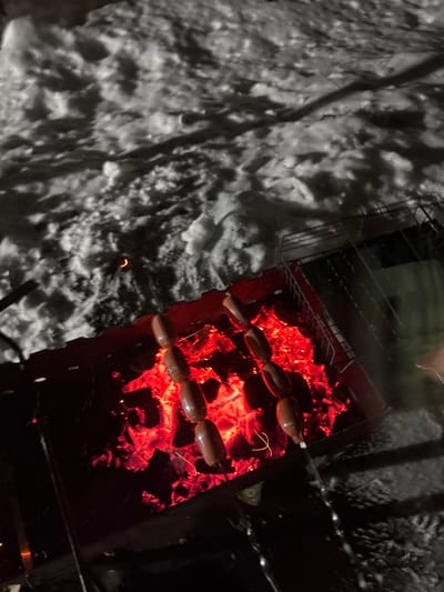 A charcoal grill glows with red-hot coals. Skewers hold rows of sausages, cooking over the intense heat. The stark contrast highlights the brightness of the grill against the darkness surrounding it. Adjacent to the grill, snow covers the ground. The scene creates an interplay between the warmth of the cooking fire and the cold, snowy environment.