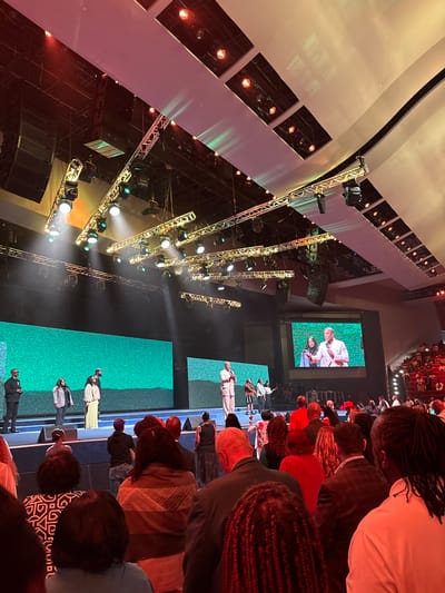 The image depicts an indoor gathering with a stage and audience. On stage, several individuals are visible, with two prominent figures speaking to the audience, one of whom is also displayed on a large screen behind them. The audience is seated in rows of red chairs, facing the stage. The environment appears to be a large venue, possibly a church or convention center, with a complex lighting and sound system suspended from the ceiling. The overall atmosphere suggests a public event, potentially a service or presentation. The specific location in Dallas, United States of America, is not evident from the image itself.