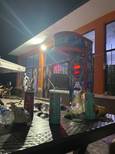 The image depicts an outdoor scene at night, likely in Tri Kladentsi, Bulgaria, centered around a boxing arcade machine. A young man in a white and grey t-shirt and grey shorts stands near a bright orange building with large windows. To the left, a white umbrella shades a seating area where other individuals are seated. The foreground is dominated by a table laden with various items including empty plastic bottles, cans of what appears to be energy drink and iced tea, and some food packaging. The boxing machine, illuminated by spotlights, features a digital display showing "0000" for score and "2978" for high score. The environment suggests a casual gathering or entertainment spot, possibly at a gas station or roadside rest stop, given the presence of the arcade machine and the general ambiance. The ground appears to be a paved or gravel surface, and the lighting is primarily artificial, casting strong shadows.