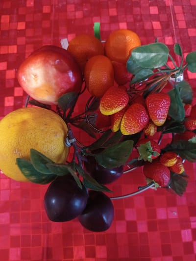 A still life arrangement of artificial fruits is displayed on a red checkered surface. The fruits include several bright red strawberries with yellow tips, a cluster of small orange tangerines, dark purple plums, a red and yellow apple, and a yellow lemon. The fruits are attached to a wire frame with artificial green leaves and brown branches. A small white label with the number "26" is visible on one of the branches. The lighting suggests an indoor setting, and there are no discernible weather or time of day cues. The overall impression is one of a decorative display rather than a real fruit selection. The location context provided is Pavlohrad, Ukraine.