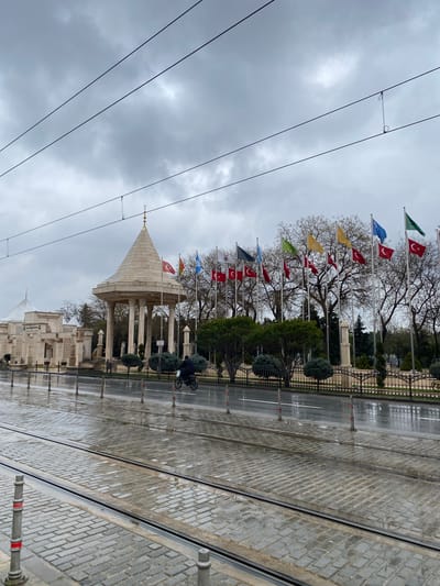 This outdoor shot depicts a street scene in Karatay, Turkey on an overcast day, oriented with the top of the photo to the left. Running diagonally across the scene from the upper left to the lower right are tram lines embedded in a cobblestone street. Along the center line of the image, a row of flagpoles with various national flags lead towards a mausoleum-style building with a cone-shaped roof. Trees with bare branches stand behind the flags. A dark-clad person on a bicycle is visible on the right, moving along the wet pavement. Electric lines are overhead, crisscrossing the cloudy sky. The overall impression is one of a quiet, possibly rainy, day in a historic area of Karatay.