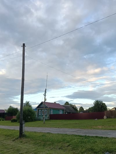 This media file depicts a rural scene in Smolino, Russia, likely during the late afternoon or early evening, given the soft, diffused light and cloudy sky. The image is dominated by a wooden utility pole in the foreground on the left, with overhead wires stretching across the frame. In the middle ground, a traditional Russian wooden house, painted turquoise with green trim and a red roof, stands behind a dark red fence. The house has decorative window frames, suggesting a common architectural style for the region. To the right of the house, another utility pole is visible, and further to the right, more houses and trees can be seen, hinting at a village or small settlement. The foreground is a grassy area bordering a dirt or gravel road. The sky is filled with dramatic, textured clouds, illuminated by what appears to be the setting sun, creating a serene and somewhat moody atmosphere. There are no people visible in the image, and no text is present. The overall impression is one of quiet, rural life in a rustic setting.