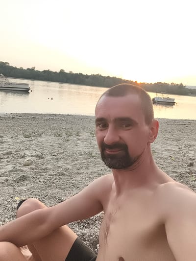 A man with a short buzz cut and a goatee is sitting on a pebble beach next to a body of water, with trees lining the opposite bank. The sky is bright and overcast, suggesting either dawn or dusk. Two boats are visible on the water: one larger pontoon boat to the left and a smaller motorboat to the right. The man is shirtless and wearing dark shorts. He is looking towards the camera, with his left leg crossed over his right. The scene appears to be a natural setting within the city of Dunakeszi, Hungary.