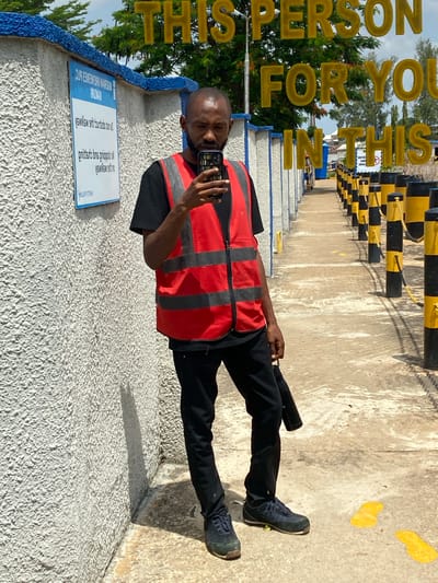 In Kinkinau, Nigeria, a Black male individual stands on a paved pathway beside a textured white wall. He is dressed in a black t-shirt, black pants, and a reflective red vest. The individual is holding a smartphone in his right hand, appearing to take a photo or video. The pathway is lined with alternating black and yellow bollards connected by yellow chains. In the background, a blue and white sign is affixed to the wall. The sky above is partly cloudy. The phrase "THIS PERSON FOR YOU IN THIS" is superimposed on the image in large yellow letters.
