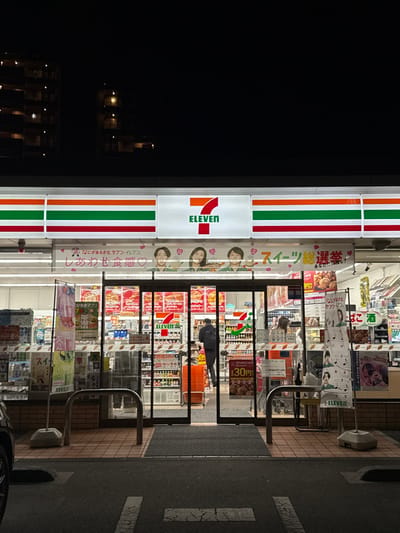 The image displays the brightly illuminated facade of a 7-Eleven convenience store at night, located in Nagano, Japan. The store's prominent red '7' and green 'ELEVEN' logo is centered above the main entrance, flanked by horizontal orange, green, and red striped panels. Below the main sign, a large banner features three stylized faces and Japanese text, including phrases such as "しあわせ食感" (Happy Texture) and "スイーツ総選挙" (Sweets Grand Election).

The entrance consists of automatic glass sliding doors. A solitary individual, seen from behind, wearing a dark jacket and backpack, is partially visible mid-stride, either entering or exiting the store. Through the extensive glass storefront, the brightly lit interior reveals rows of shelves stocked with various products, including beverages in refrigerated units on the left and numerous packaged goods. An orange shopping basket is visible near the entrance inside. Additional promotional posters and banners are affixed to the glass, some advertising "酒" (sake/alcohol) and "SEVEN CAFE" with a "30円" (30 yen) price point.

In the foreground, an asphalt parking area with faint white markings is visible. The immediate area fronting the store features light brown paved tiles and includes metal bollards or railings. The background consists of the dark silhouettes of multi-story buildings, characterized by scattered illuminated windows, under a dark night sky.