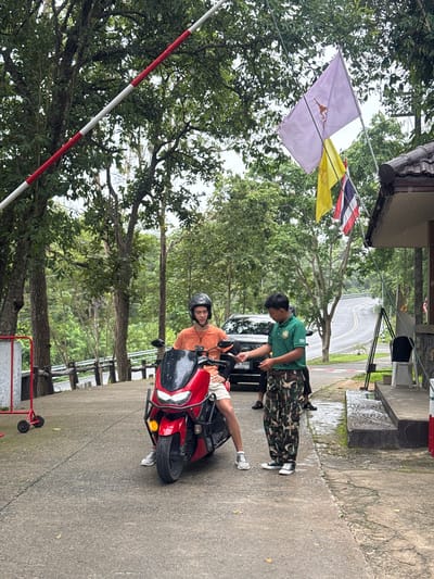 The image depicts a scene at what appears to be an entrance to a park or attraction in Suthep Town Municipality, Thailand. A young man wearing a helmet and an orange t-shirt is seated on a red scooter. He is being spoken to by another individual wearing a green t-shirt and camouflage pants, who is gesturing towards the scooter. Behind them, a black car is parked.

In the background, a winding road leads into a lush, green, forested area, suggesting a natural or park-like setting. Flags, including what appears to be a purple flag with an emblem and the Thai national flag, are visible, indicating a potentially official or significant location. A red and white striped barrier is raised overhead, suggesting a checkpoint or entrance gate. The ground is paved and appears wet, possibly from recent rain. The overall environment is characterized by dense foliage and a humid, tropical atmosphere.