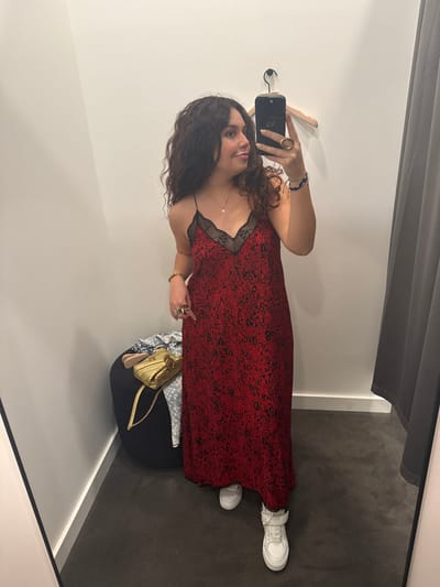 A woman in a red dress with black lace trim and white sneakers stands in a dressing room. She holds a phone to take a selfie. A hanger is mounted on the wall, and a black curtain hangs from the ceiling. A handbag sits on a chair. The walls of the dressing room are white and the floor is grey.