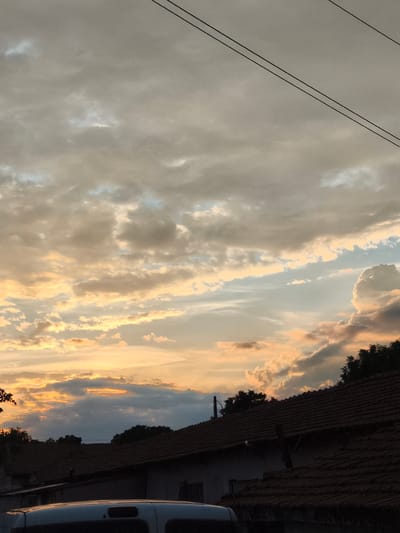 The image shows a view of the sky above rooftops in Orizari, Bulgaria. The sky is filled with clouds, some of which are illuminated by the setting sun with hues of orange and yellow. There are also power lines visible in the upper part of the image. The foreground features the rooftops of buildings, some with dark tiled roofs, and a portion of a white vehicle. The scene appears to be a residential area.