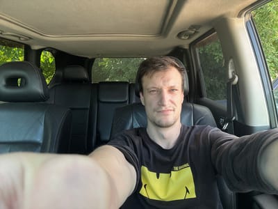 The image is a selfie taken by a man sitting in the driver's seat of a car. He is wearing a black t-shirt with a yellow graphic and the words "THE ORIGINAL" visible on it. The interior of the car is visible, including black leather seats and a sunroof. Outside the car, lush green trees are visible, suggesting an outdoor setting. The lighting indicates it is daytime, likely late afternoon due to the angle of the sun. The man's expression is neutral, and he appears to be looking directly at the camera. The background includes the back seats of the car and windows with a view of foliage. The overall scene suggests the man is either parked or stopped during a drive. Based on the provided location context, the scene is set in Shatava, Ukraine.