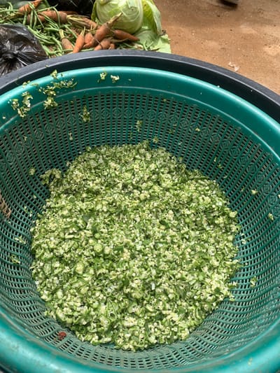 In Zhwarki Mile 10, Nigeria, a close-up shot captures a green plastic basket filled with chopped okra. The okra pieces are small and irregularly shaped, revealing their pale inner seeds and green outer skins. The basket itself has a patterned texture with numerous small holes, allowing for drainage. In the background, out of focus, are other vegetables including carrots, green beans, and a head of cabbage, along with a black plastic bag. The ground appears to be sandy. The lighting suggests it is daytime, likely outdoors.