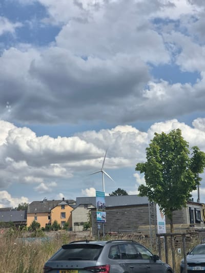 This image depicts a cloudy sky with a wind turbine visible in the distance, suggesting an outdoor setting in a rural or semi-rural area. A lush green tree is prominent in the foreground, casting shadows. Below the tree, a residential area with several houses can be seen, one painted orange and another in a neutral tone. A dark-colored car is parked in the foreground on the right side of the image, with a license plate that reads "MM 7601". The weather appears to be partly cloudy, with patches of blue sky interspersed with large, fluffy white clouds and some darker, more ominous clouds. The overall impression is that of a somewhat overcast day, possibly late afternoon or early evening, given the lighting. There are signs visible near the houses and tree, though the text is too small to be legible in the provided image. The scene appears peaceful and suburban, with no people immediately visible. The presence of the wind turbine and the houses suggests a location that combines elements of nature, renewable energy, and residential living.