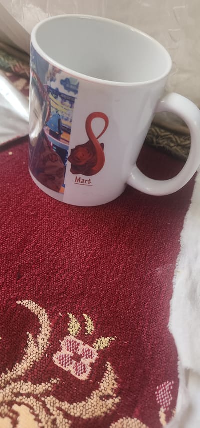 The image shows a white ceramic mug with a printed design. The design features a red rose superimposed on a stylized red number 8, with the word "Mart" printed below it. To the left of the rose and number 8, there is a partial image of a person's face and upper body, with colorful background elements. The mug is placed on a textured, dark red surface with a repeating pattern. The background is blurred, suggesting a shallow depth of field.

It is not possible to determine the location in the city of Türkmenbaşy, Turkmenistan from the provided image.