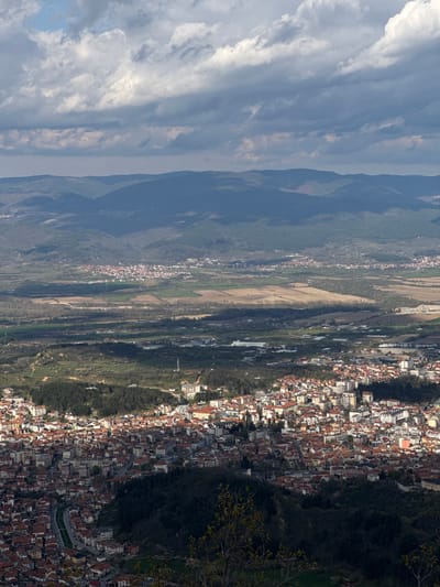 The image presents a high-angle landscape view of Delchevo, Bulgaria. The scene encompasses a valley transitioning into a dense urban area. The sky is characterized by numerous clouds. Mountains with green, tree-covered slopes are on the left and behind the city. A mix of fields and smaller settlements are visible in the valley between the mountains and the main urban area.
The city itself is a collection of tightly packed buildings, predominantly with red rooftops. Vegetation is interspersed within the urban layout, with trees and grassy areas. A winding road runs along the edge of the city near some trees on the right.