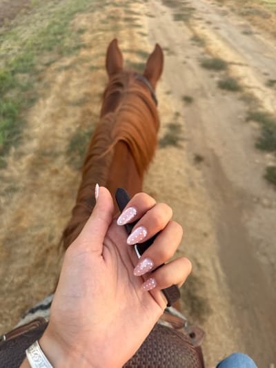 The image depicts a first-person view of a person riding a horse on a dirt trail in Rancho Murieta, United States. In the foreground, a human left hand with manicured, almond-shaped nails, painted light pink with white polka dots, grips a dark strap, likely a rein. A silver bracelet with an embossed pattern is visible on the wrist. Below the hand, a portion of a brown saddle or saddle blanket is present, along with a small visible section of blue denim apparel. The midground shows the back and prominent mane of a chestnut-colored horse, with its ears pointed forward and a dark bridle strap visible around its head. The background consists of a dusty, unpaved trail showing tracks, bordered by sparse, dry golden and green vegetation. The scene is illuminated by natural light.
