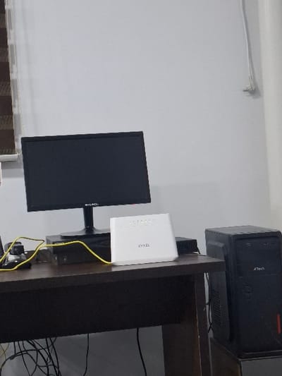 The scene is an indoor computer workstation located in Kumaryk, Uzbekistan. A dark brown desk occupies the mid-ground, positioned against a light-colored, likely white, wall. On the desk, a black Everich flat-screen monitor with an unlit screen is centrally placed. To the right of the monitor, a white ZYXEL router/modem is present, displaying several illuminated green indicator lights. A dark, rectangular electronic device, possibly a set-top box or DVD player, is situated beneath the monitor stand. A bright yellow cable connects to the router and extends to the left. To the right of the desk, a black A Tech desktop computer tower, featuring a red vertical accent stripe on its lower front panel, stands on an adjacent surface. A tangled collection of black cables is visible beneath the desk. On the far left, a section of horizontal blinds with brown and white slats is partially visible. A white cable runs vertically along the right wall. No individuals are present, and no active interactions are depicted.