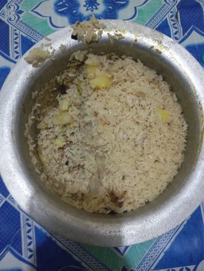 An aluminum cooking pot, circular in form, is depicted containing cooked rice. The rice is predominantly off-white to light brown, mixed with visible yellowish chunks identified as potatoes and darker fragments, likely caramelized onions or spices. The pot's interior is filled approximately two-thirds with the food. The rim of the pot exhibits minor wear. The pot rests on a patterned surface, likely a tablecloth, featuring repeating geometric and floral designs in shades of blue, white, and light green. No individuals, actions, or interactions are visible in the scene. The location is Nkomo ward, Kenya.