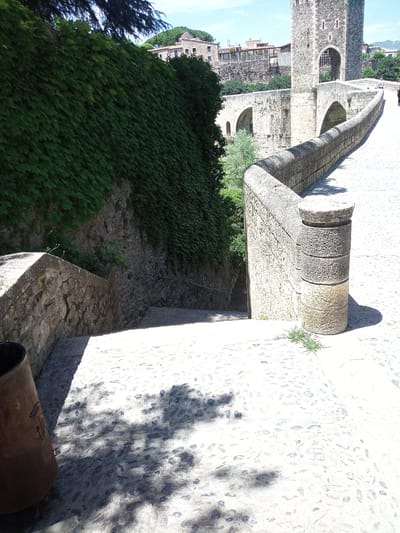 The image depicts the medieval bridge of Besalú, Spain. The foreground features a cobblestone pathway leading to a stone staircase, bordered by a low stone wall covered in ivy on the left. To the right, a rounded stone pillar with stacked segments marks the edge of the bridge. In the background, the stone bridge continues, featuring an arched passageway and a stone tower with battlements. Buildings with terracotta roofs are visible beyond the bridge, nestled within the town. The sky is clear and blue, suggesting a sunny day. There are no visible people or animals in the frame. The overall impression is of a historic and picturesque architectural setting.