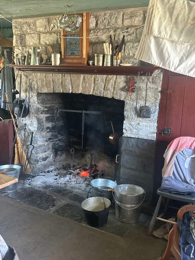 This image depicts a rustic kitchen scene, likely in an older home in South Dundas, Canada. The focal point is a large, stone fireplace where a fire is burning, indicated by glowing embers and rising smoke. Several metal buckets, some appearing to be galvanized, are positioned around the fireplace, suggesting tasks related to fire maintenance or water storage.

The kitchen area is filled with various objects that evoke a historical or traditional setting. On the left side of the fireplace, there are shelves and holders containing what appear to be cooking utensils, brooms, and possibly brushes. Above the fireplace, a red wooden cabinet door is visible, with a white cloth hanging down on the left. A chair draped with fabric is placed near the fireplace on the right.

A smaller framed picture or tile with a blue and gold pattern is mounted within a wooden frame above the fireplace, adding a decorative element to the stone wall. The overall atmosphere is one of a working kitchen from a past era, with the presence of the fire and an assortment of tools and containers contributing to this impression. There is no visible text in the image. The time of day and weather are not discernible from the image.