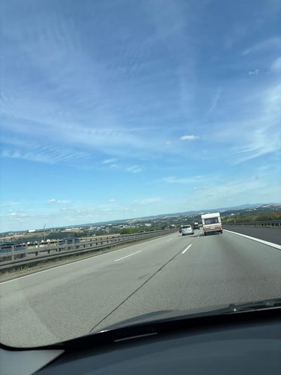 The image shows a view from inside a vehicle traveling on a highway in Elz, Germany. The sky is blue with scattered white clouds. A highway stretches ahead, with several vehicles visible, including a white caravan being towed by a car. To the left of the highway, there is a metal guardrail and beyond that, a landscape with buildings, trees, and hills in the distance. The road appears to be well-maintained.