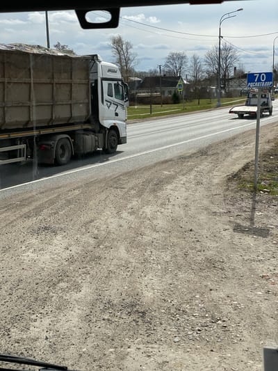 The image shows a road in Bol'shoe Teshkovo, Russia. A large white truck carrying a load of concrete blocks is driving on the road. A speed limit sign indicating "70" in Russian is visible along the road. The surrounding environment consists of trees, buildings, and a dirt shoulder next to the road. The image appears to be taken from inside a vehicle, possibly another truck.