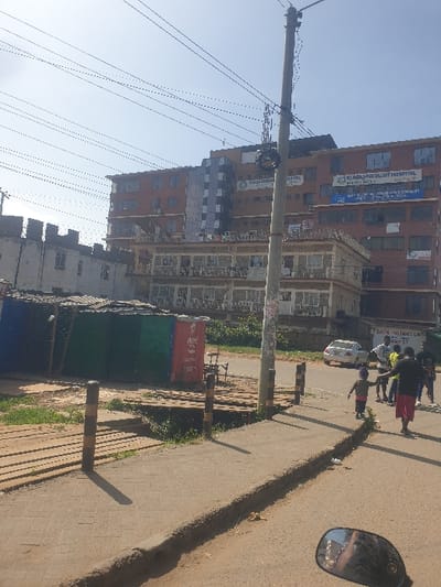 A street scene features a cluster of buildings, including Plaza Specialist Hospital. A utility pole with numerous wires stands prominently. Several people walk along the road's edge, near trash bins. The scene is captured from a high angle, possibly from a vehicle, as indicated by a side mirror visible in the right corner. The road shows signs of wear, and the overall lighting suggests a bright, sunny day.