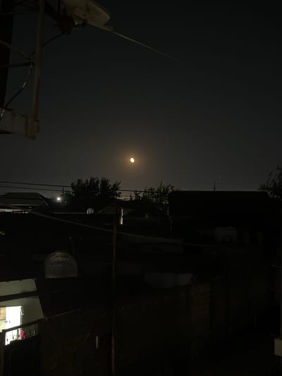 The image depicts a nighttime view over rooftops in the city of Sabunchu, Azerbaijan. The sky is dark, with a bright, partially illuminated moon visible in the upper right quadrant. Silhouetted against the sky are several trees and the dark shapes of buildings, some with sloped roofs. Power lines are strung across the scene, and a water tank with the label "TURKSAN" is visible on one of the rooftops. A portion of a metal structure with an attached light fixture is in the upper left corner. Some windows of buildings show interior light. The overall impression is a dimly lit urban landscape at night.