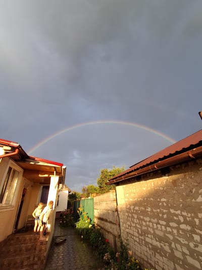 The image captures a residential yard in Krolevets, Ukraine, under a dramatic sky featuring a rainbow. Two young boys stand on the steps of a house with a covered porch, their attention seemingly directed towards something in the distance. The house has light-colored walls and a dark red, sloped roof. Adjacent to the house is a paved pathway leading between it and a weathered brick wall. Green foliage and colorful flowers are visible along the pathway and bordering the wall, suggesting a garden area. A green metal fence runs along the right side of the yard. The sky is overcast with dark clouds, but a portion of a rainbow is clearly visible arching across the frame, indicating recent rainfall. The sunlight casts a warm glow on the side of the house and the brick wall, contrasting with the darker sky. A scooter lies on the paved path. The overall scene depicts a moment of natural beauty following or coinciding with a rain shower, observed in a private outdoor space.
